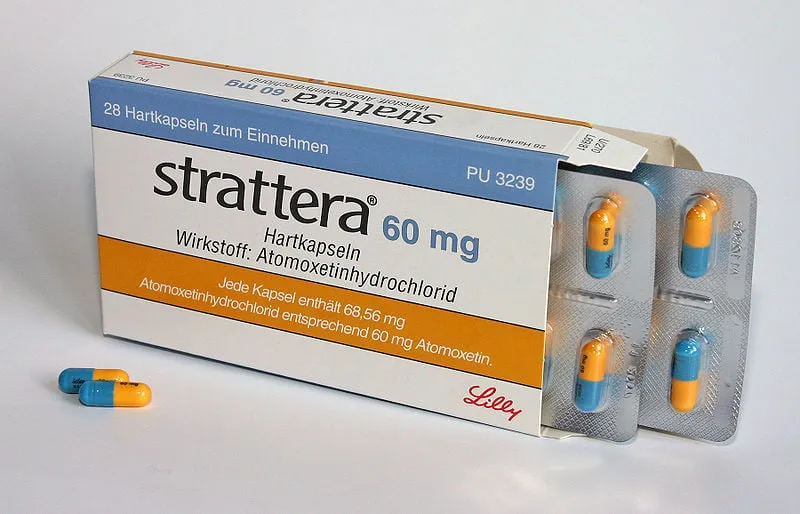 Atomoxetine A box of Strattera (Atomoxetine) 60 mg hard capsules, showing the German packaging with "Hartkapseln" and "Wirkstoff: Atomoxetinhydrochlorid," along with two blue and orange capsules outside the blister packs, indicating medication for ADHD.