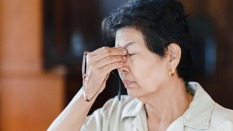 A senior Asian woman, possibly with cataracts, is holding her eyeglasses and rubbing her nose bridge/forehead due to eye strain, blurred vision, or eye pain. She appears to be squinting or experiencing discomfort from glare/bright light.