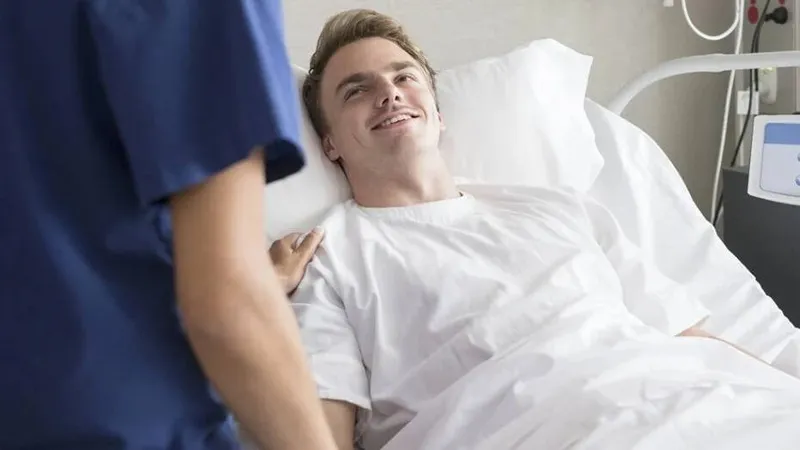 A smiling young man in a white hospital gown lying in a hospital bed, receiving reassuring support from a healthcare professional (nurse or doctor) wearing a blue uniform, emphasizing patient care during or after treatment.