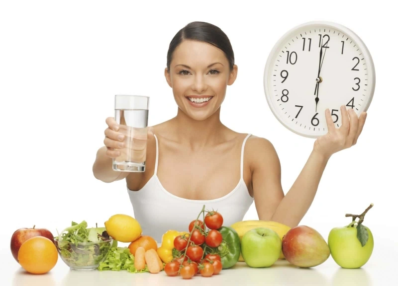 Voice Disorders A woman holding a glass of water and a clock, with a spread of fruits and vegetables, illustrating a healthy diet and hydration for voice disorder prevention