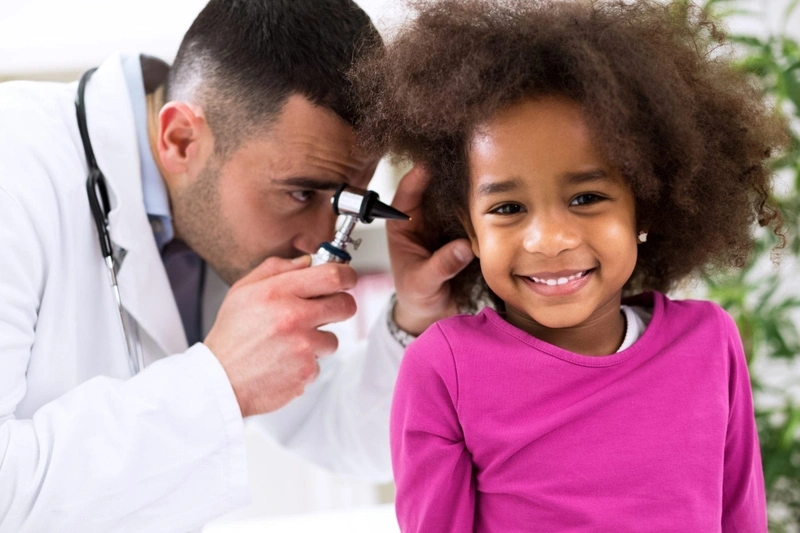 A pediatrician using an otoscope to examine the ear of a smiling young girl during a medical checkup