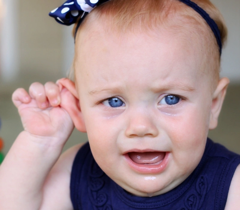 A young toddler girl crying and tugging her ear, showing common signs of an ear infection or fluid buildup in children